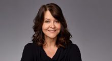 A woman with brown hair and a black scoop next top smiling against a grey backdrop