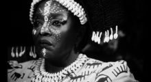 An African woman frowns, she dons elaborate face paint and shells woven into her hair. She is shot via black and white photography.