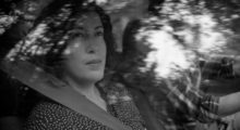 A black and white image of a woman sitting in the passenger seat of a car looking out the window. She has shoulder-length wavy hair and wears a blouse with small polka dots on it.