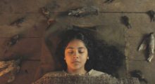 A woman with long, black hair sleeps on a cot with a burlap blanket.