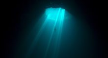 A freediver ascends through an undersea cave as a bright blue sunbeam shines down on them.