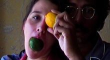 A man and a woman stand next to each other. The woman holds a lime in her mouth and places a lemon over her left eye. The man wears glasses and his face is semi-obscured by her head and arm.