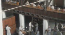 Monks in long, white robes work on watches in a multi-level building embellished with dark wood fixtures.
