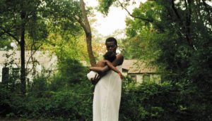 A person wearing an off-white long tunic holds a baby in their arms. They are surrounded by lush, green trees and a semi-obscured house.