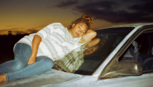A pregnant woman wearing a white and gray striped t-shirt and blue jeans lounges atop the hood of a car. Her brown hair is tied up in a messy bun.