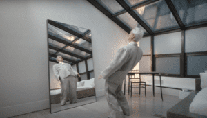David Byrne re-dons his iconic gray, oversized suit and tries it on anew in his bedroom and looks in the mirror, leaning back.