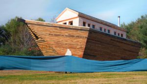 A giant, wooden arc sits in the middle of a green field. A large blue tarp simulates the look of water below it.