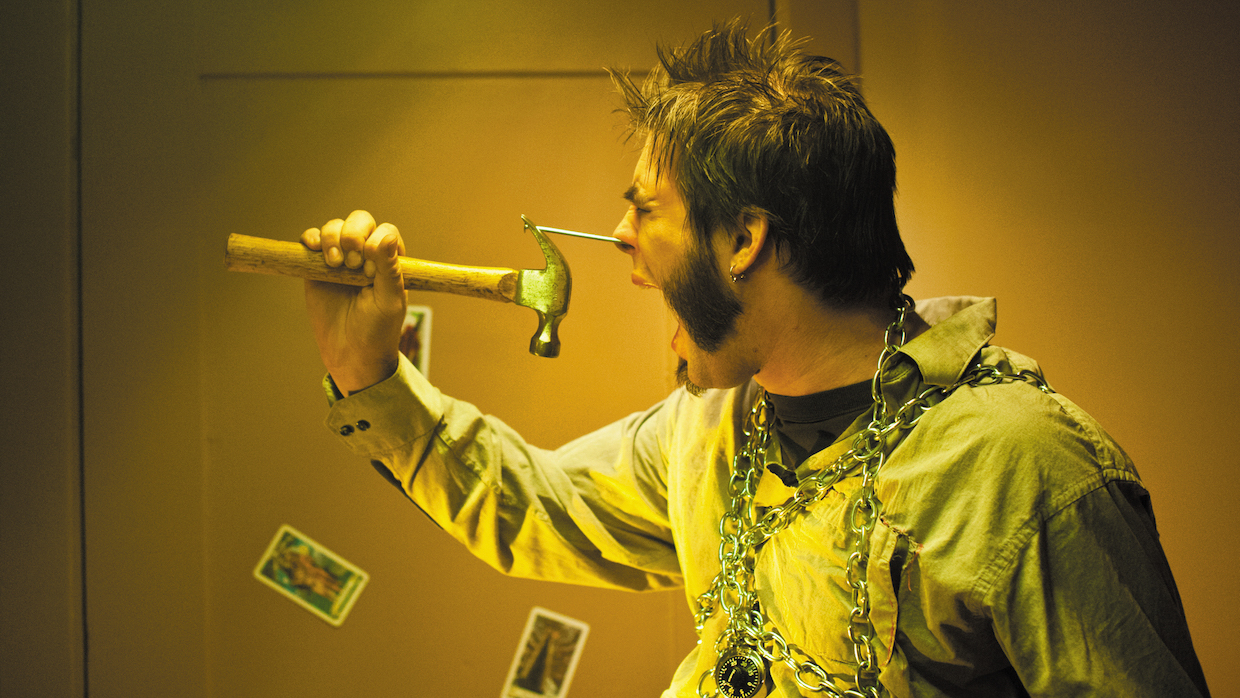 A man with a spiky faux-hawk and exaggerated sideburns appears in anguish as he sticks a nail in his nose and wears a chain around his torso secured by a padlock.