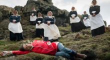 A woman in a red raincoat and blue jeans lies on her side on a rocky moor; she is surrounded by foreboding Puritan women.