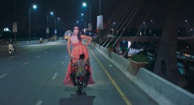 A man rides his motorcycle on an open highway, with a cardboard cutout of a woman in traditional Pakistani garb attached to the front of his vehicle.