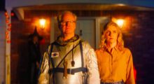 A middle-aged man wears an astronaut suit and stands next to a younger woman wearing a salmon colored button-up shirt.