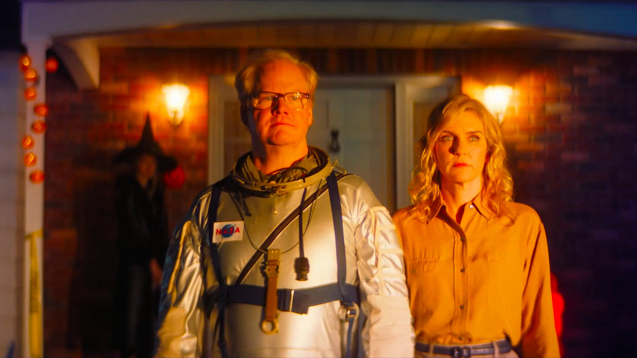 A middle-aged man wears an astronaut suit and stands next to a younger woman wearing a salmon colored button-up shirt.