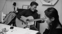 A man wearing a black turtleneck plays guitar for a young woman who looks off into the distance.