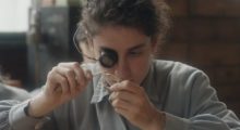 A young woman with curly brown hair pulled back examines a watch with a magnifying glass and tweezers.