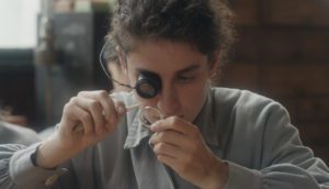 A young woman with curly brown hair pulled back examines a watch with a magnifying glass and tweezers.