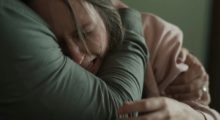 A woman with long, dirty blond hair sobs as a man in a long-sleeve gray shirt holds her head to his chest. We cannot see the man's face.
