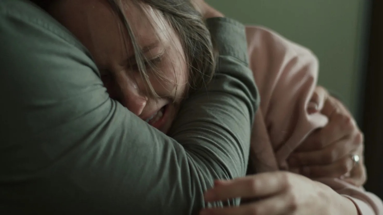 A woman with long, dirty blond hair sobs as a man in a long-sleeve gray shirt holds her head to his chest. We cannot see the man's face.