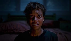 A young Black woman with cropped short hair sits up in a dark room, her eyes widen as she gazes at something that clearly terrifies her.