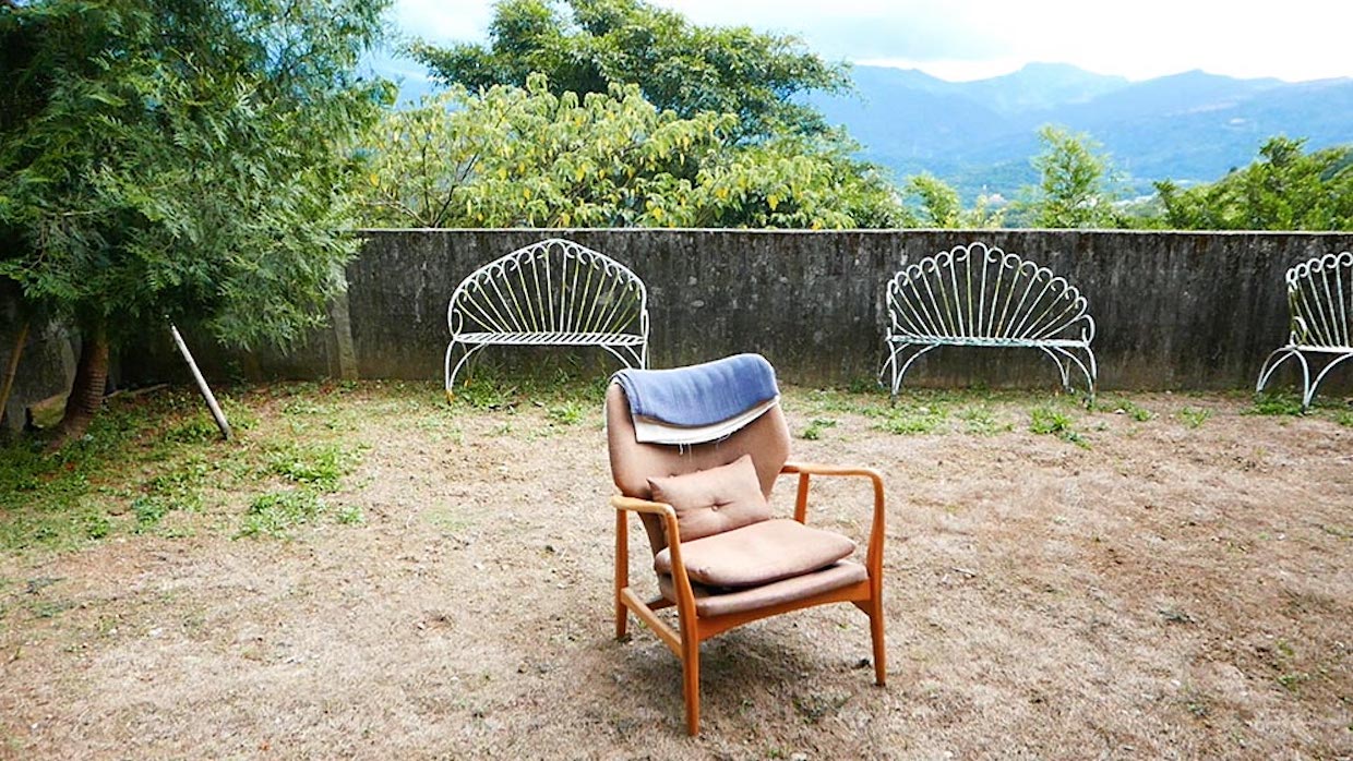 An empty wooden chair sits in a yard, the grass barely grows and is surrounded by a stone wall.