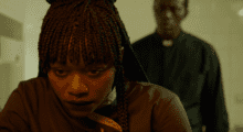 A close-up shot of a Black woman with braided hair. She leans over a counter while a priest looms over her from behind.