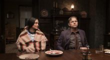 A woman wearing an orange and tan hand-woven shawl sits at a dining table next to a man in a dark-toned suit. She looks at him and he looks away into the distance. The room is like by a few stray gas lamps and the plates they have eaten from are empty.