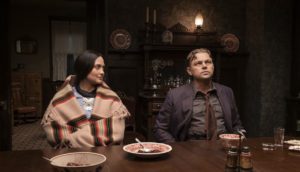 A woman wearing an orange and tan hand-woven shawl sits at a dining table next to a man in a dark-toned suit. She looks at him and he looks away into the distance. The room is like by a few stray gas lamps and the plates they have eaten from are empty.