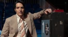 A man wearing a '70s era tan suit and orange-hued tie stands with his arm propped up against a vintage TV camera.