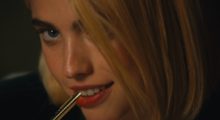 A close-up shot of a woman in a blond wig smiling as she bites down on a pen.