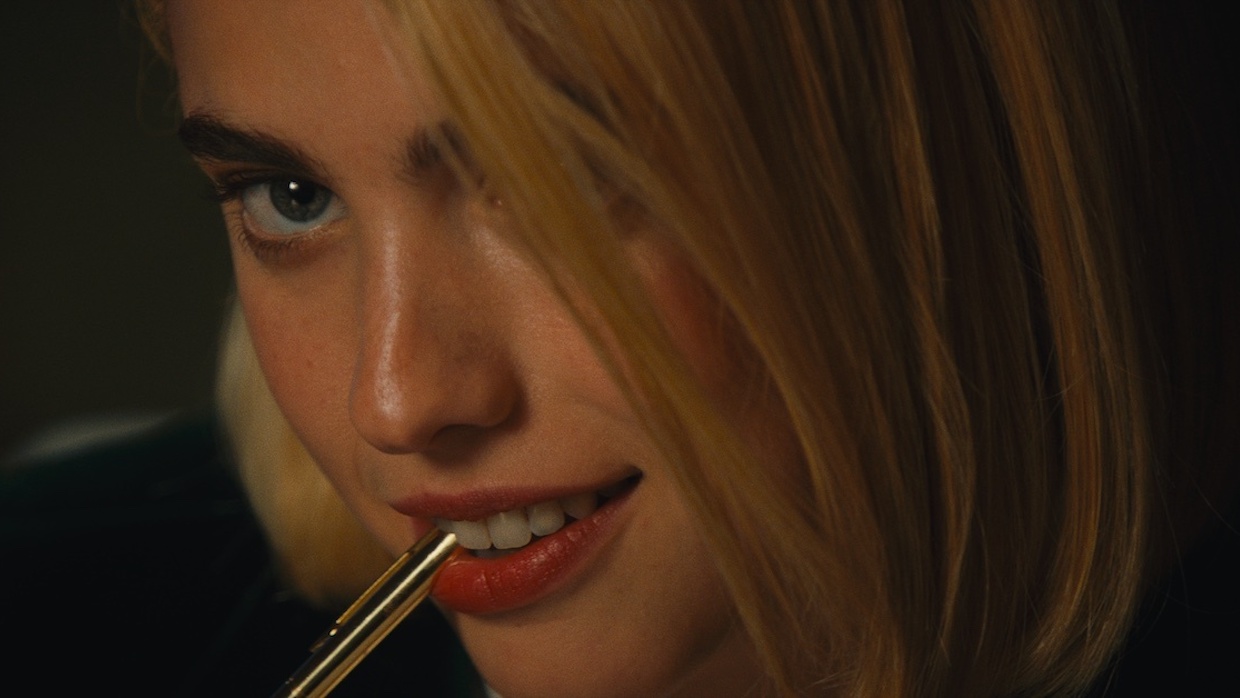 A close-up shot of a woman in a blond wig smiling as she bites down on a pen.