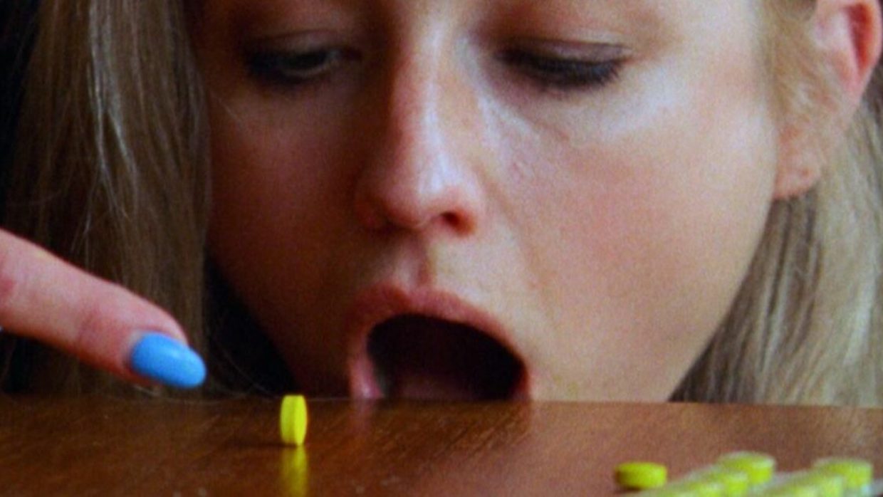 A blond woman with neon blue painted nails opens her mount near the edge of a table and rolls a bright green pill into her mouth.