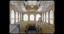 A woman sits on an empty old-fashioned trolley, she sits and stares straight ahead and the windows let in flat, white light.