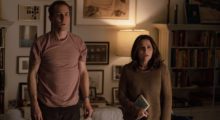 A man and a woman stand in casual clothes in their apartment. They both look at something with shock on their faces.