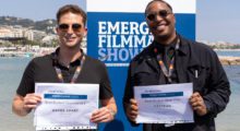 Two young men stand in front of the Emerging Filmmaker Showcase at Cannes and hold up paper certificates that show they've won.