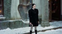 A woman with long black hair stands on a snowy sidewalk in front of a gray stone building with eccentric oblong windows. She wears a black peacoat with exaggerated shoulder pads and gray thigh-high socks with black knee-high boots.