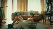 A woman sits nude in a plastic washtub in the middle of her living room. Her extremities also soak in similar plastic buckets.