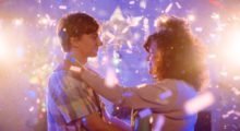 Bright pink and purple lights wash over a man and a woman dancing at what appears to be a prom.