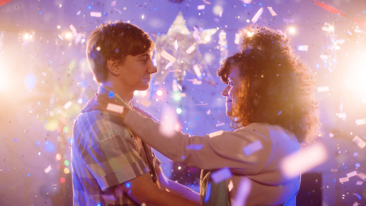 Bright pink and purple lights wash over a man and a woman dancing at what appears to be a prom.