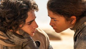 A woman (Zendaya) in futuristic protective sci-fi attire caresses the face of a man (Timothée Chalamet) amid a barren desert landscape.
