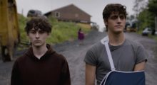 A boy with cropped curly hair and a brown hoodie stands next to his older brother, who's a bit taller and wears a gray t-shirt with his arm in a sling. In between them but behind in the distance is their mother, whose outline is blurred by her distance.