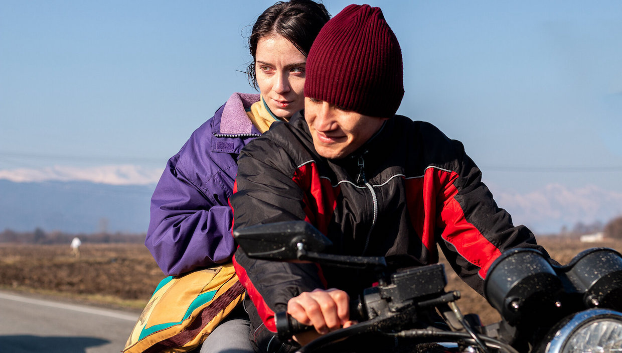 A woman and a man wear light jackets. The man drives a motorcycle and the woman sits behind him on the vehicle.