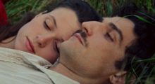 A young couple lie in the grass together. The woman rests her head on the man's shoulder.
