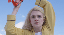 A young women with platinum blond hair holds something out of sight above her head as she gazes in front of her. She is wearing a yellow long-sleeve button-up top.