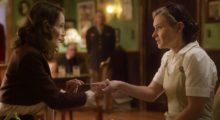 Two women sit in a '40s-era style parlor room, one woman wearing a military uniform is holding a long needle against the finger of the other woman, who is blonde and wears beige overalls and a white linen shirt.