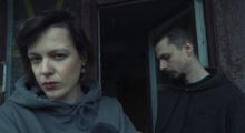 A man and a woman stand next to each other. The woman faces forward and looks directly at the camera, while the man stands slightly behind her and gazes downward, only showing his side profile.