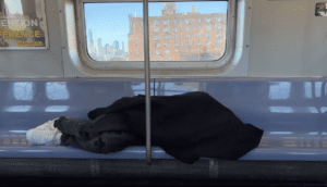 A man sleeps with a jacket covering his face and body on a vacant row of subway seats, Lower East Side housing projects appear outside of the window behind him.