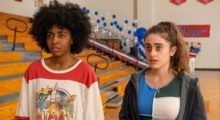 Two girls stand next to each other in a high school gymnasium. On the left is a Black girl with an afro wearing a red and white t-shirt. Standing next to her is a white girl with curly, medium-length hair wearing a patchwork shirt with different hues of blues and whites.