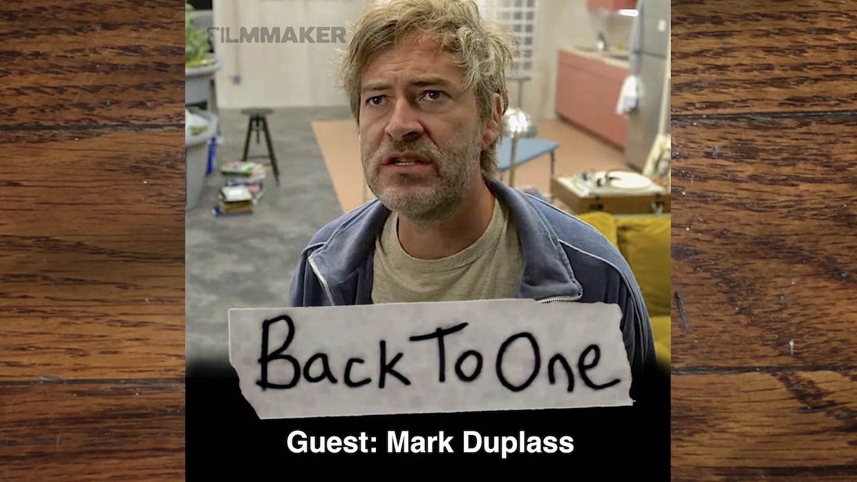 An image of actor and filmmaker Mark Duplass, who wears a beige t-shirt, grey hoodie and scowls while looking off to the side.