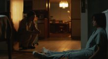 A woman wearing blue hospital scrubs sits on a kitchen floor with her back against a wall and her feet extended long in front of her. Another woman sits on a chair and leans on her elbows toward the other woman.