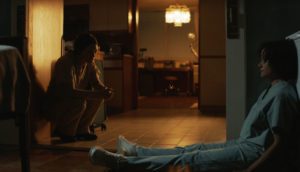 A woman wearing blue hospital scrubs sits on a kitchen floor with her back against a wall and her feet extended long in front of her. Another woman sits on a chair and leans on her elbows toward the other woman.