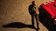 A man wearing a brown suit walks beside a bright red Ferrari.